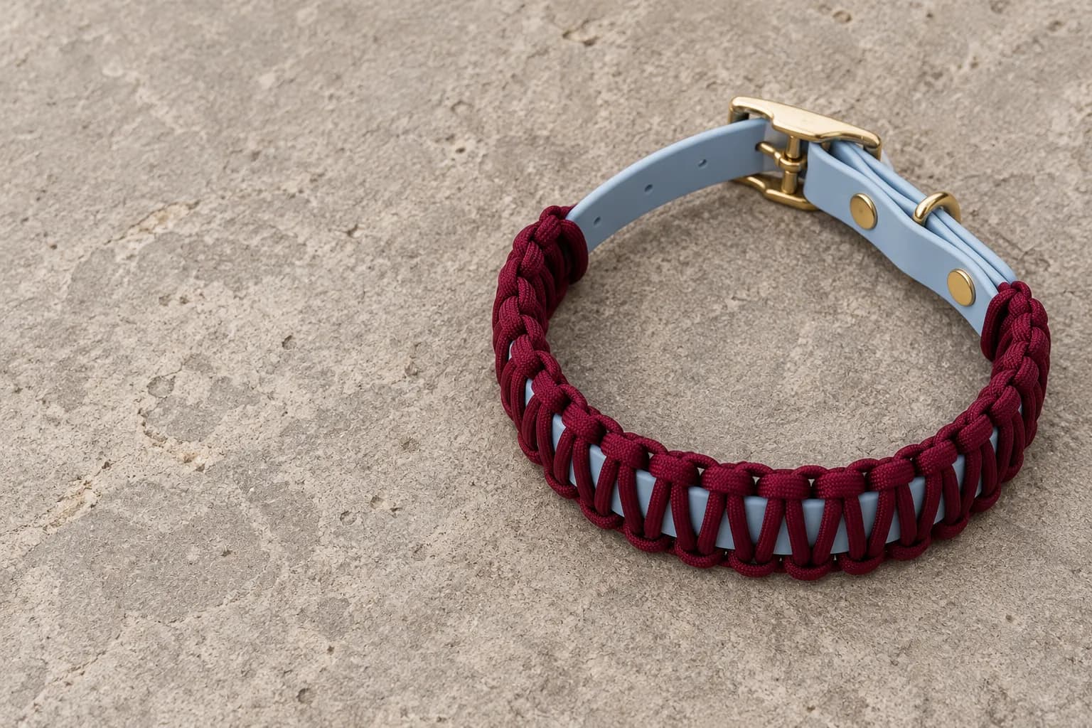 Collar with bordeaux braided cord over a light blue band and a brass buckle, on a natural stone surface.